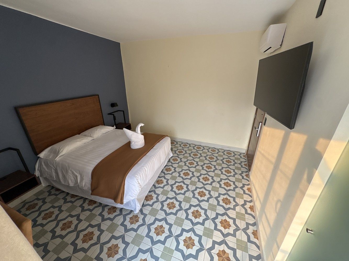 Habitación pequeña con cama, televisor montado en la pared y suelo de baldosas estampadas, bañada por la luz del sol.