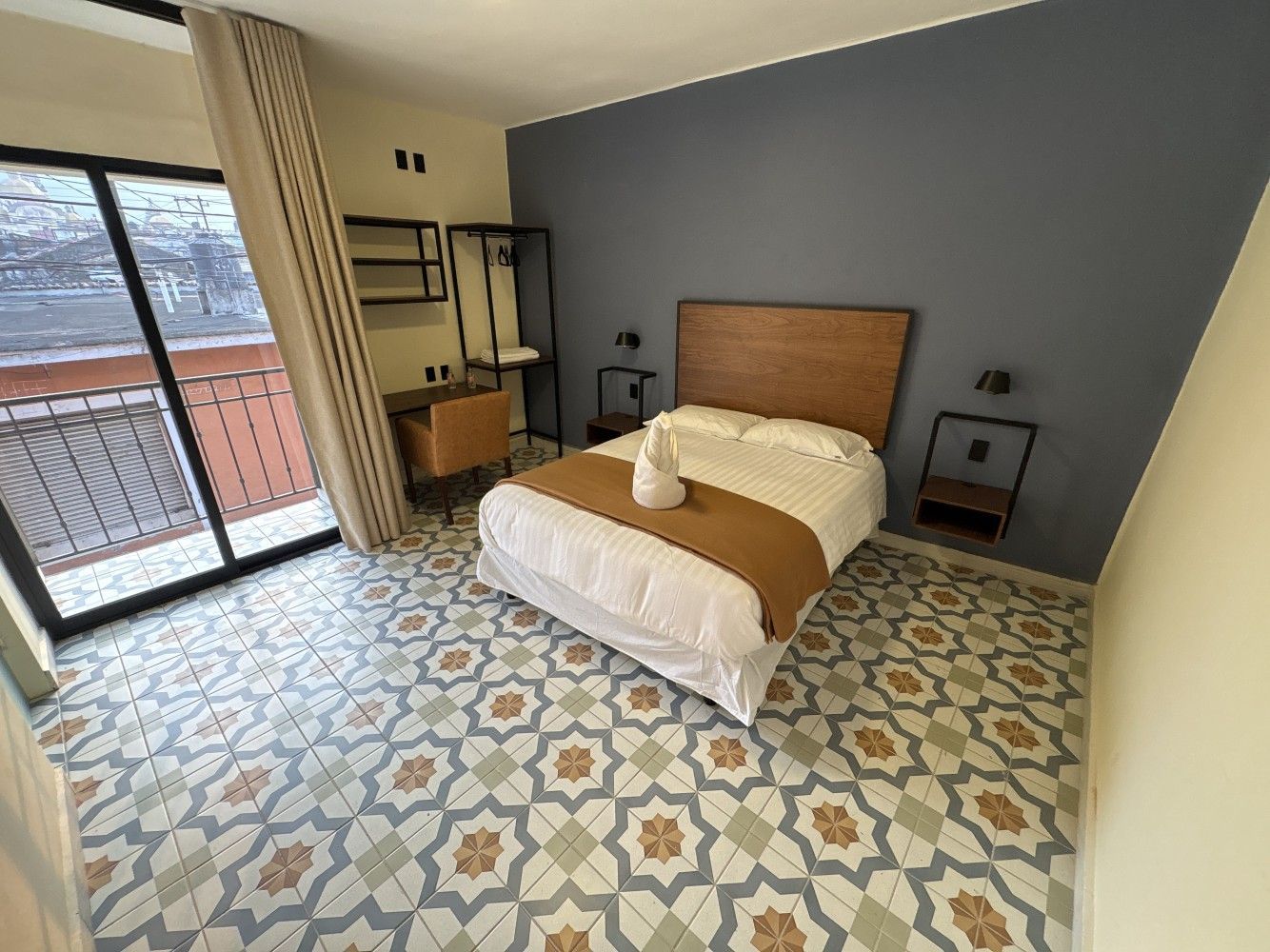 Habitación luminosa con cama, cabecero de madera, suelo de baldosas estampadas, escritorio, estanterías y puertas que dan al balcón.
