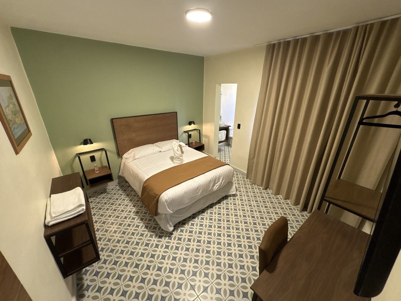 Habitación pequeña con cama, alfombra estampada, mesita de noche, escritorio y cortinas grandes.