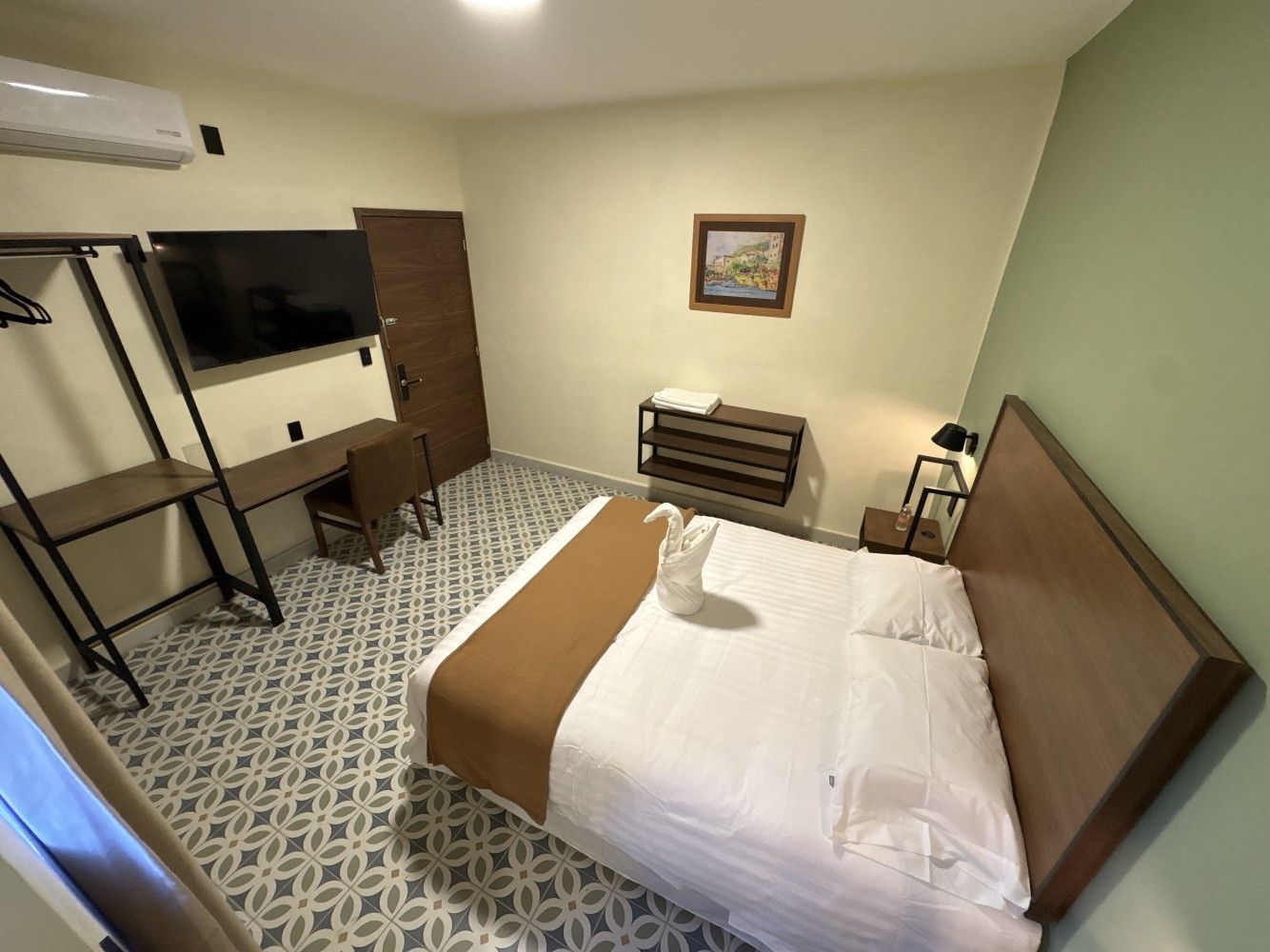 Habitación de hotel pequeña con cama, escritorio, televisor, silla, cómoda y suelo estampado.