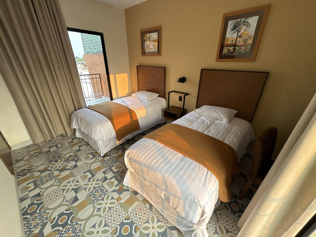 Habitación pequeña con dos camas individuales, alfombras naranjas, suelo estampado y cortinas junto a la ventana.