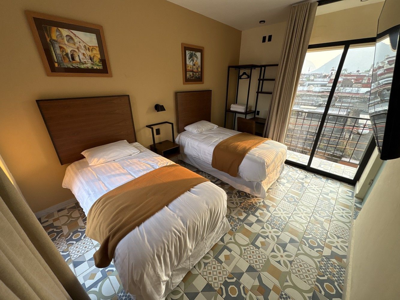 Habitación de hotel pequeña con dos camas individuales, suelo estampado, obras de arte en las paredes y balcón con vistas a través de puertas correderas.