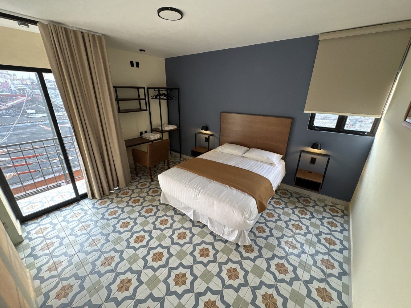 Habitación pequeña con cama, suelo de baldosas estampadas, pared decorativa azul y vistas a la ciudad desde una puerta corredera de cristal.