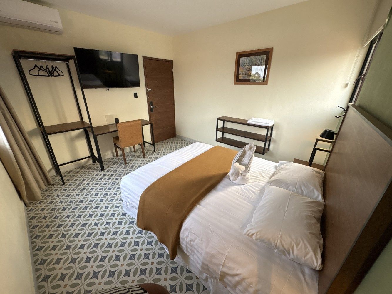 Habitación pequeña con cama, televisor montado en la pared, escritorio, silla y baldosas estampadas en el suelo.