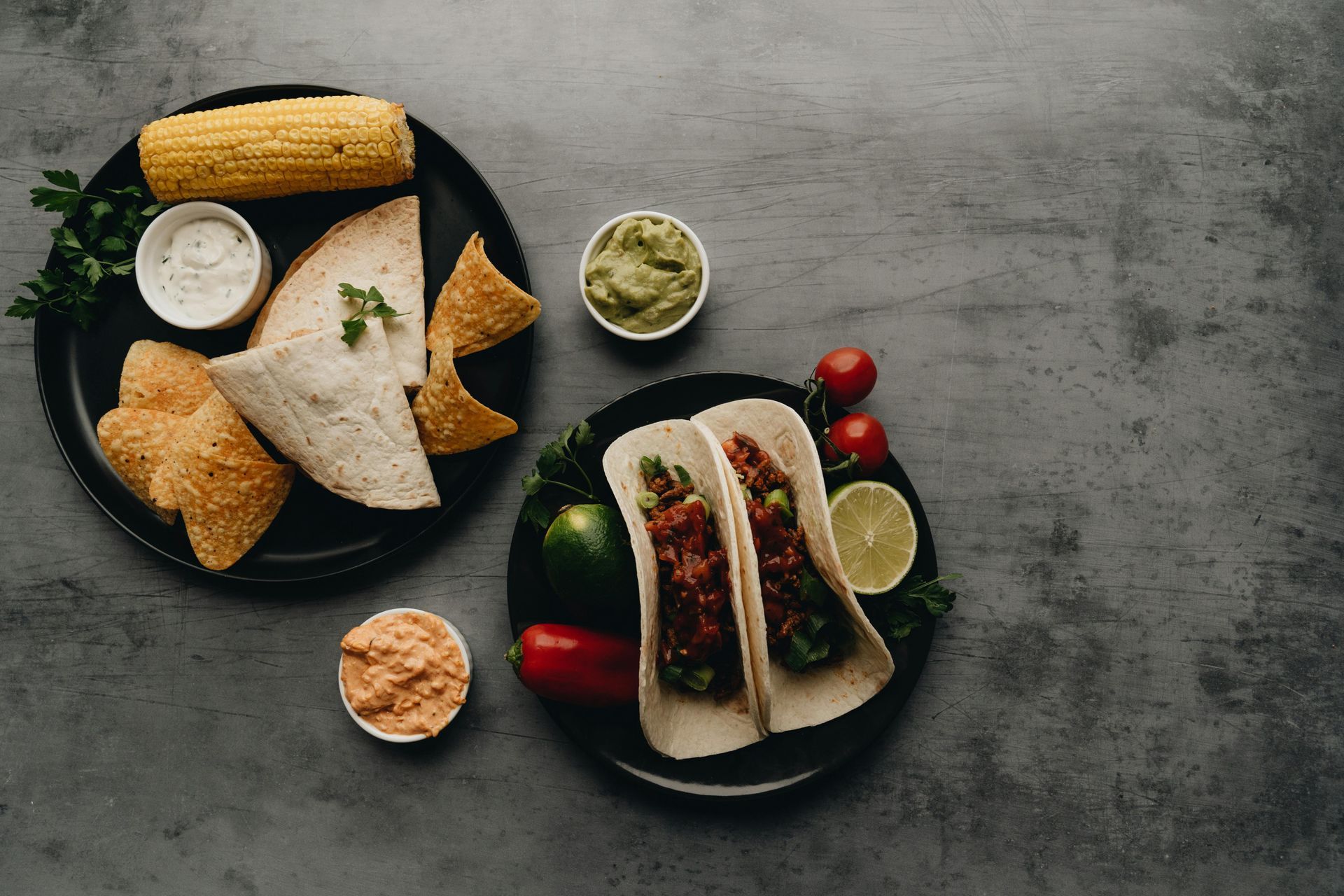 Desayuno completo con tacos, pan, salsas, maíz, lima y fresas en platos oscuros.