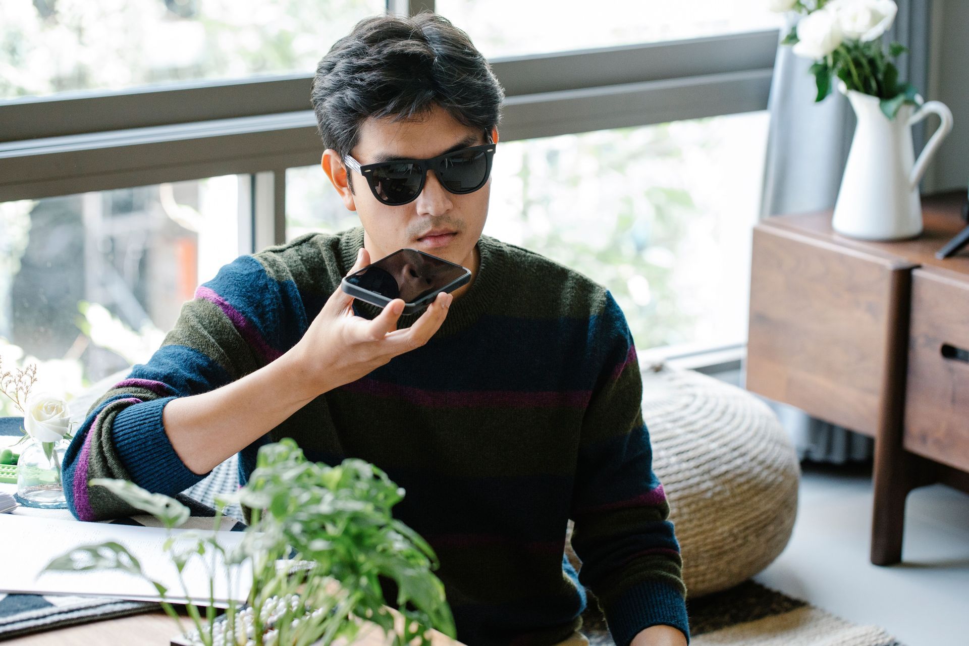 Una persona con gafas de sol habla por teléfono en una habitación luminosa con plantas y una ventana.