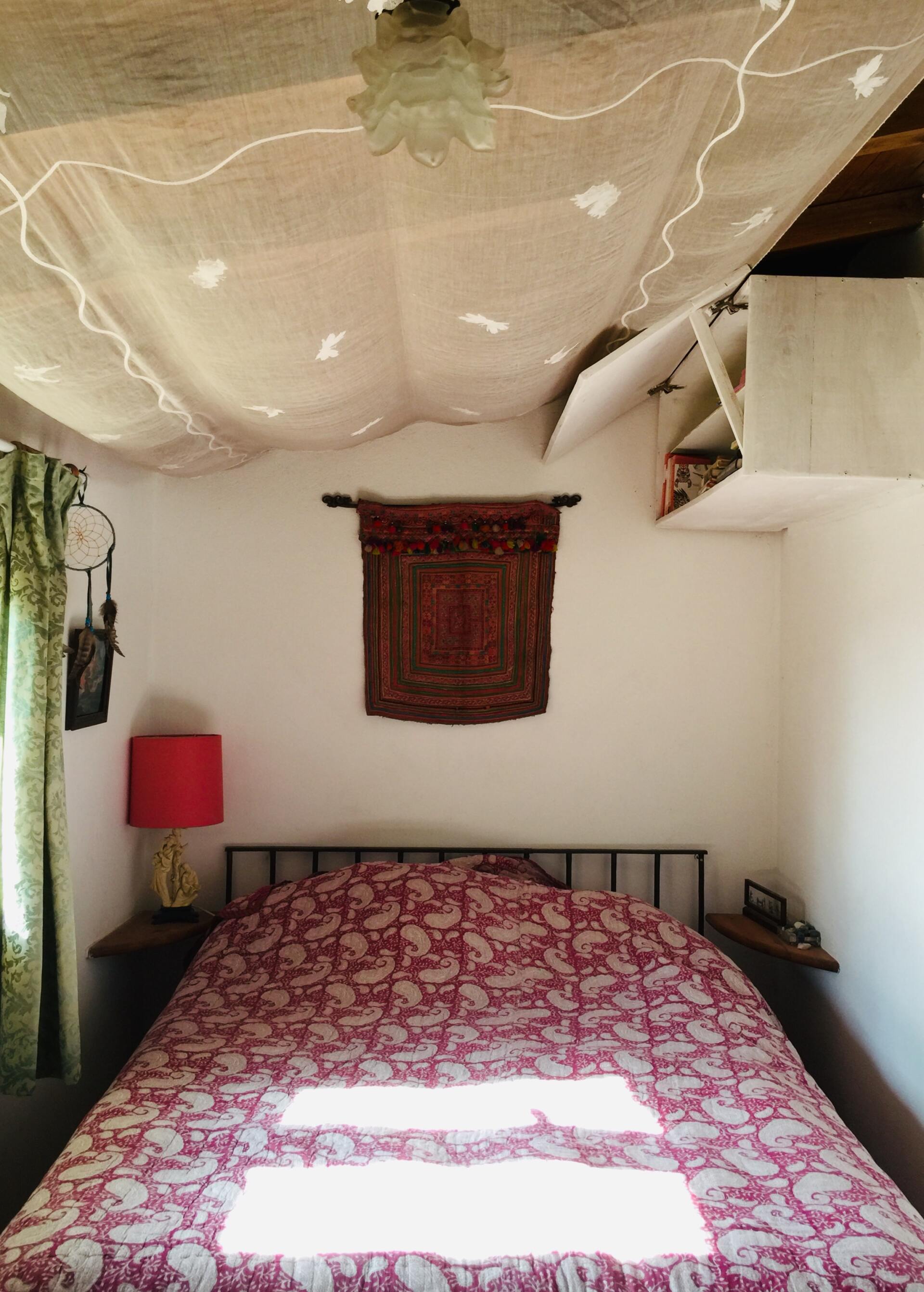 Habitación pequeña con una cama cubierta con una manta rosa estampada, una lámpara roja y un tapiz de tela en la pared.
