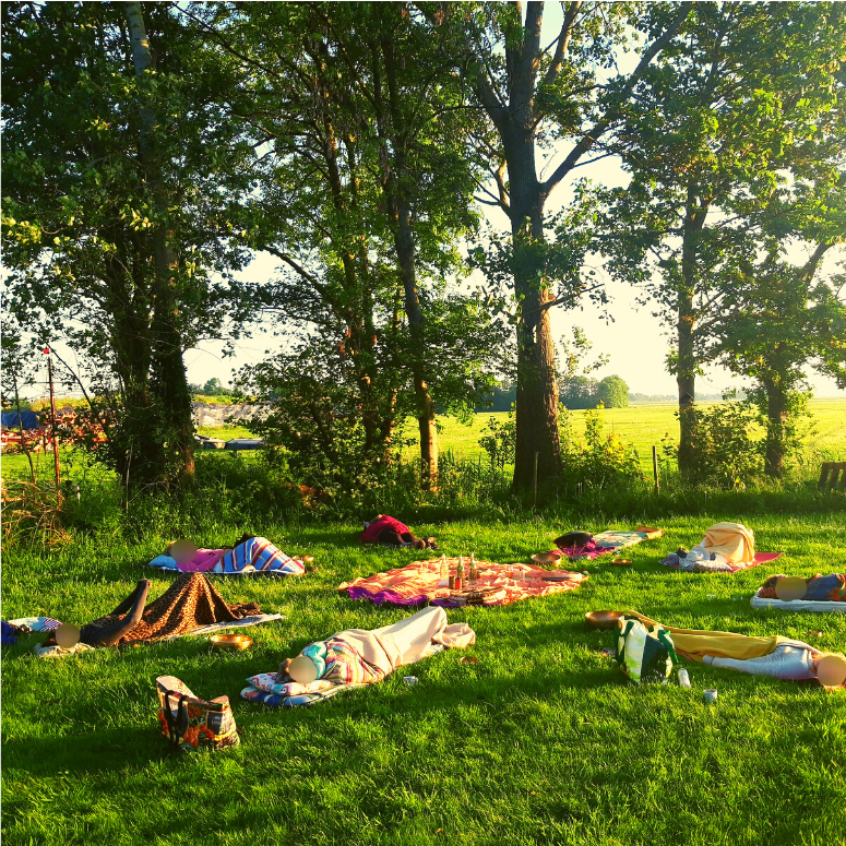 Mensen rusten op dekens in een grasveld, met bomen op de achtergrond, op een zonnige dag.