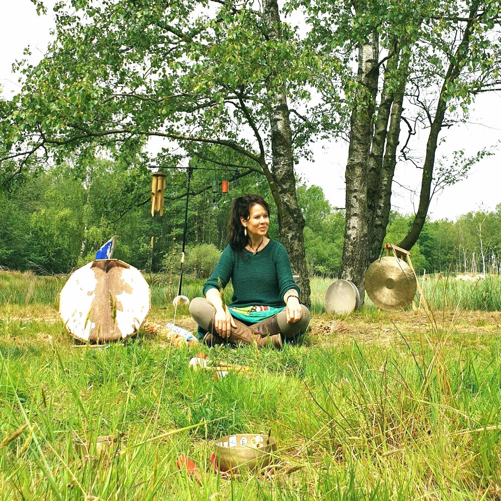 Vrouw zit in het gras onder bomen met gongs en een klankschaal.