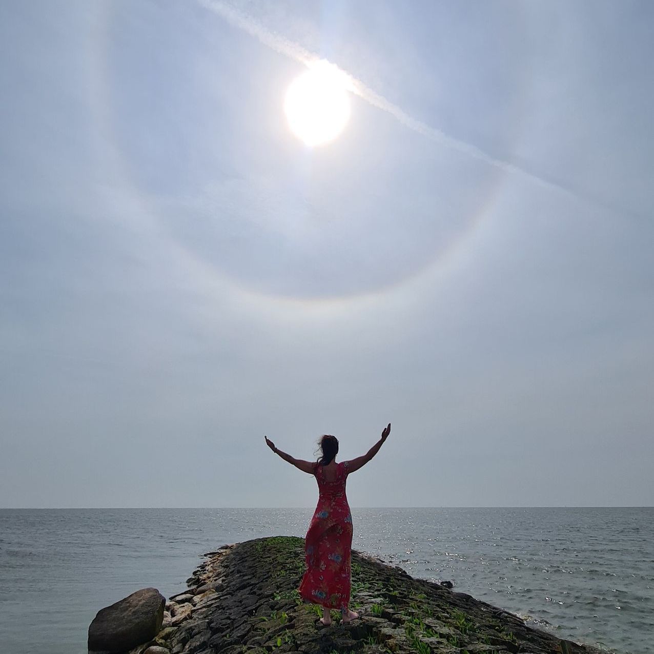 Vrouw in rode jurk met opgeheven armen, staand op een rotsachtige pier, zon met halo erboven, oceaan.