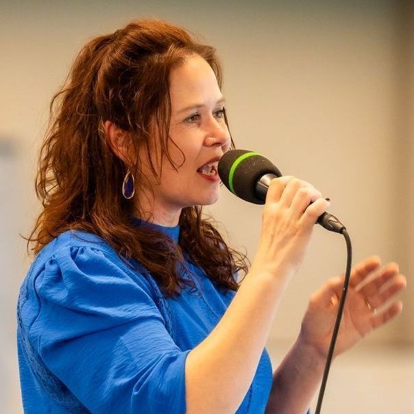 Vrouw in blauwe blouse spreekt in een microfoon, binnen.