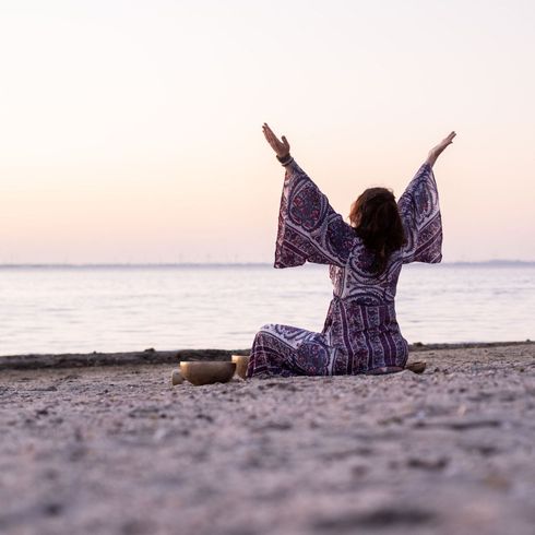 Vrouw in paarse mantel met opgeheven armen, mediterend op een strand bij zonsondergang.
