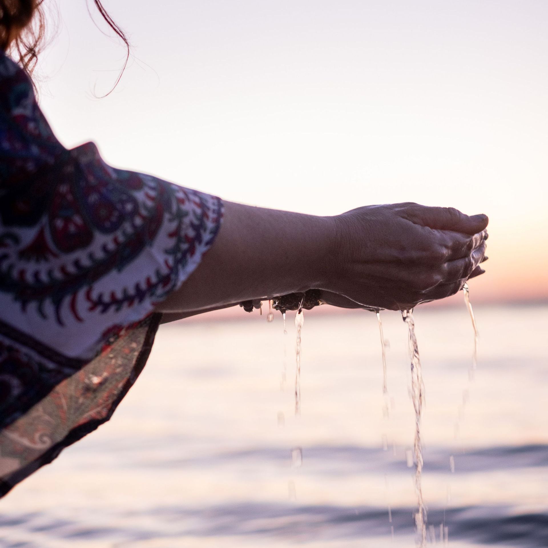 Vrouw die water in komvormige handen houdt, waar het water vanaf druppelt, bij zonsondergang boven de oceaan.