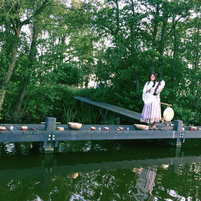 Een persoon staat op een steiger boven het water met schalen en een trommel. Bomen op de achtergrond.