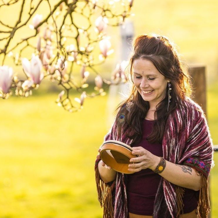Een lachende vrouw met een tamboerijn in haar handen staat buiten, vlakbij een bloeiende boom; zonnige, warme tinten.