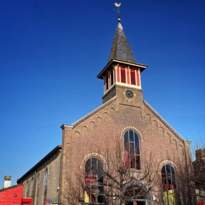 Een bakstenen kerk met torenspits, boogvensters en een windwijzer tegen een blauwe hemel.