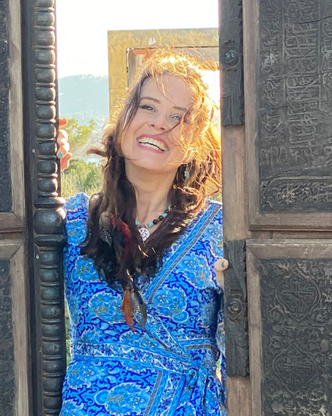 Een vrouw in een blauwe jurk met bloemenprint lacht breeduit, omlijst door een houten deuropening.