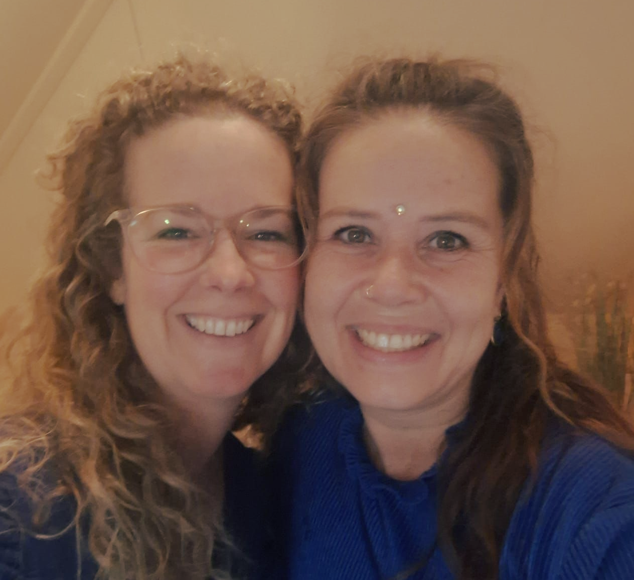 Twee lachende vrouwen, de ene met krullend haar en een bril, de andere met een neusring, in een warme binnenruimte.
