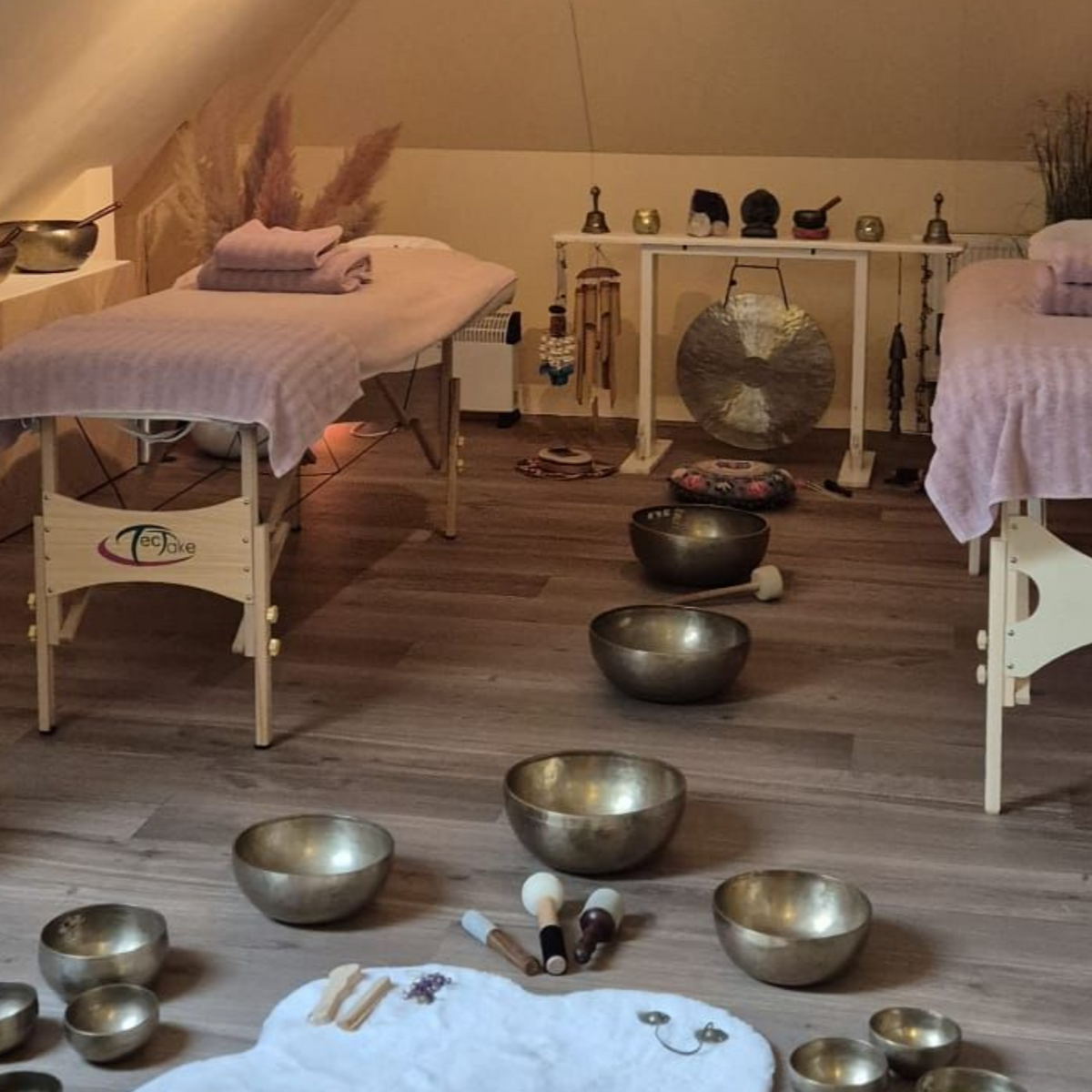 Kamer met massagetafels, klankschalen, gong en ontspanningshulpmiddelen, houten vloeren en zachte verlichting.