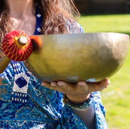 Persoon met een gouden klankschaal en hamer. Blauwe kleding met patroon, groene achtergrond.