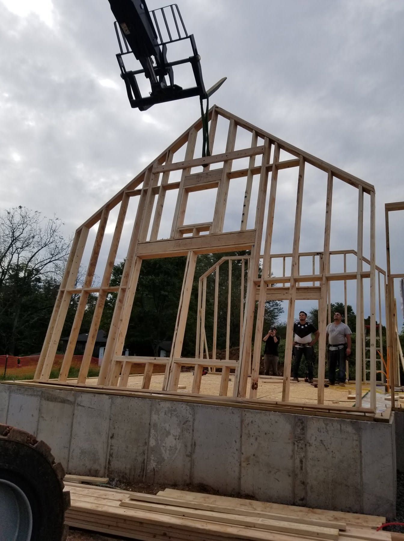 Wooden house frame under construction on a concrete foundation with workers nearby and a crane overhead
