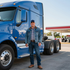 Man standing beside a blue semi-truck at a gas station, wearing a denim jacket and cap.