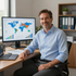 Man in a light blue shirt smiling at a desk with laptop and world map dashboard on monitor