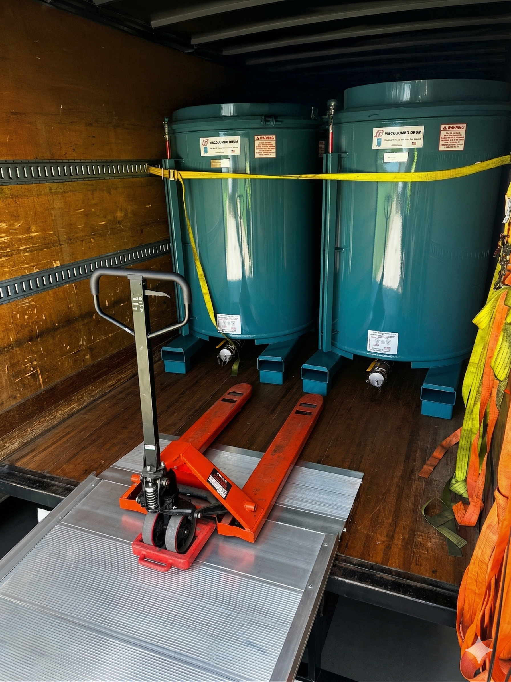 Two large teal bins strapped in a cargo truck, with a red pallet jack on the floor.