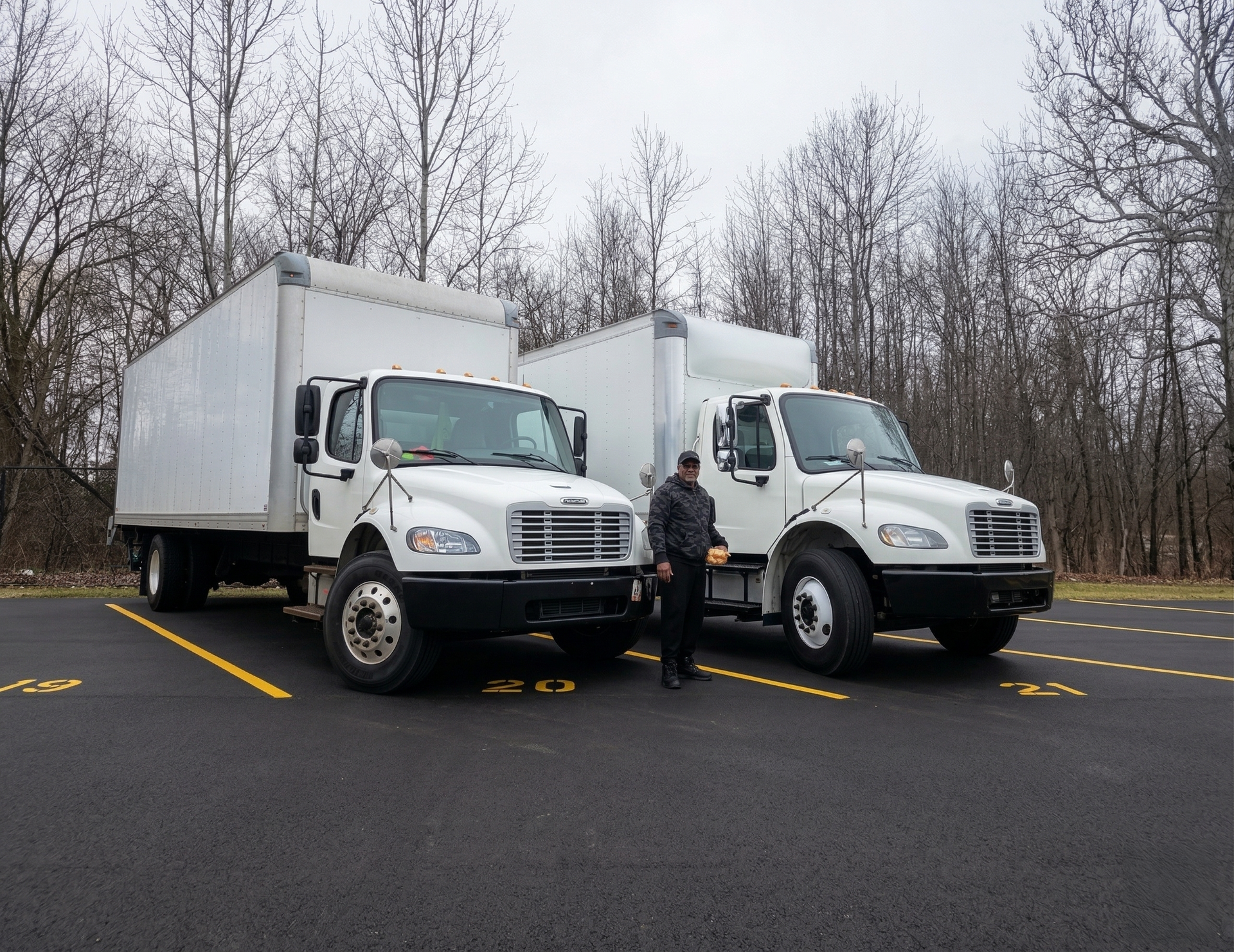 Two white box trucks parked in a lot with a person standing between them, leafless trees behind.
