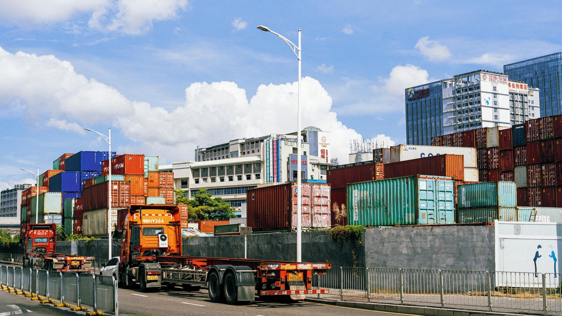 A truck pulling an empty trailer stands in front of a shipping container yard, with urban buildings visible in the back.