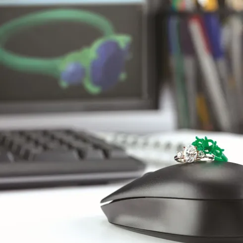 Black computer mouse with green ring and silver-green decoration on desk, keyboard and monitor blurred behind
