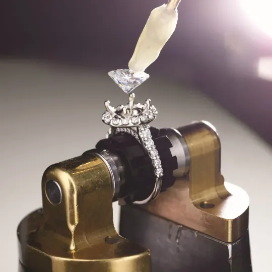 Diamond ring held in a jeweler’s clamp under a polishing tool, close-up on gold and black metal.