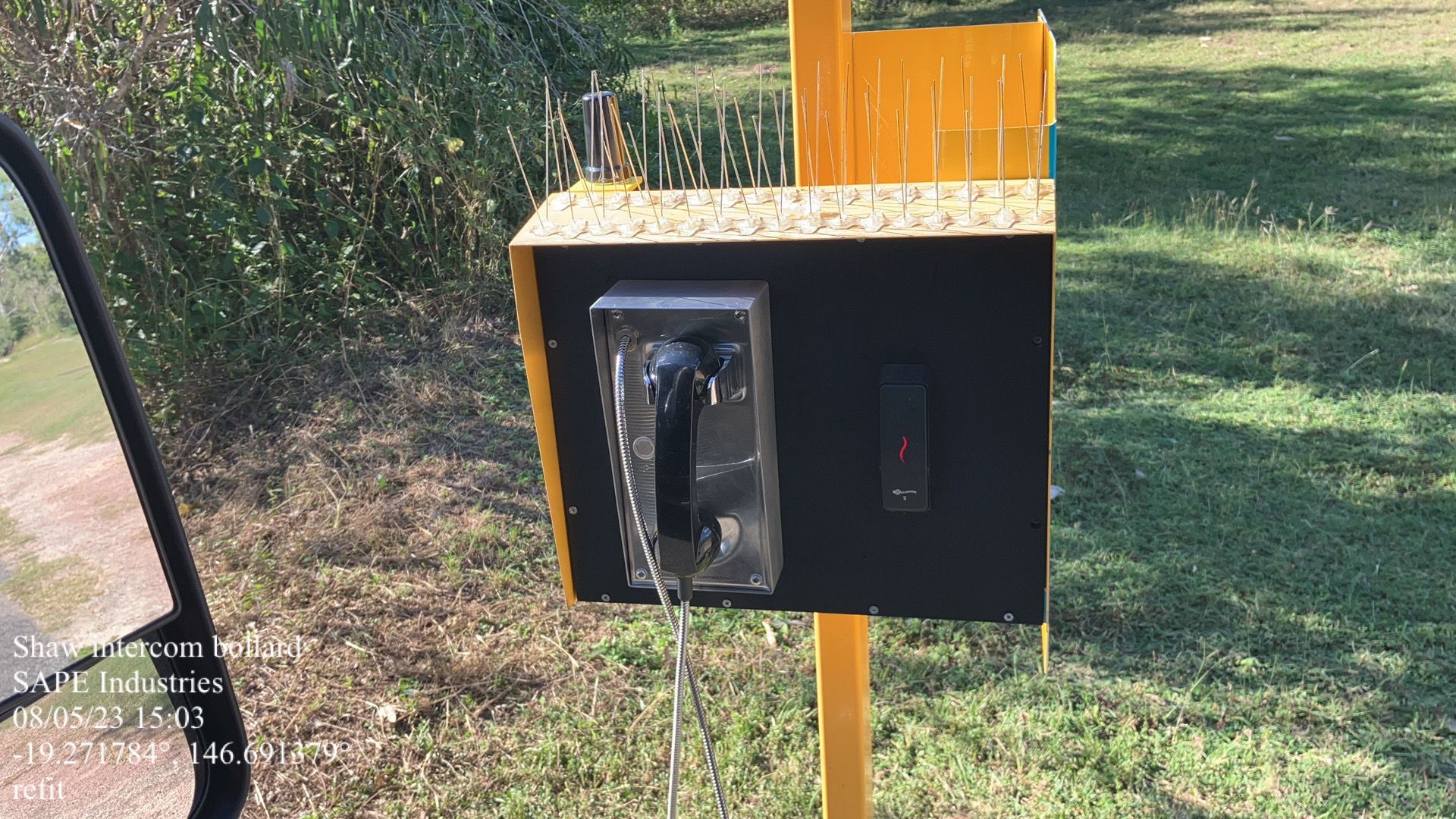 A pay phone is sitting on a yellow pole in the grass.