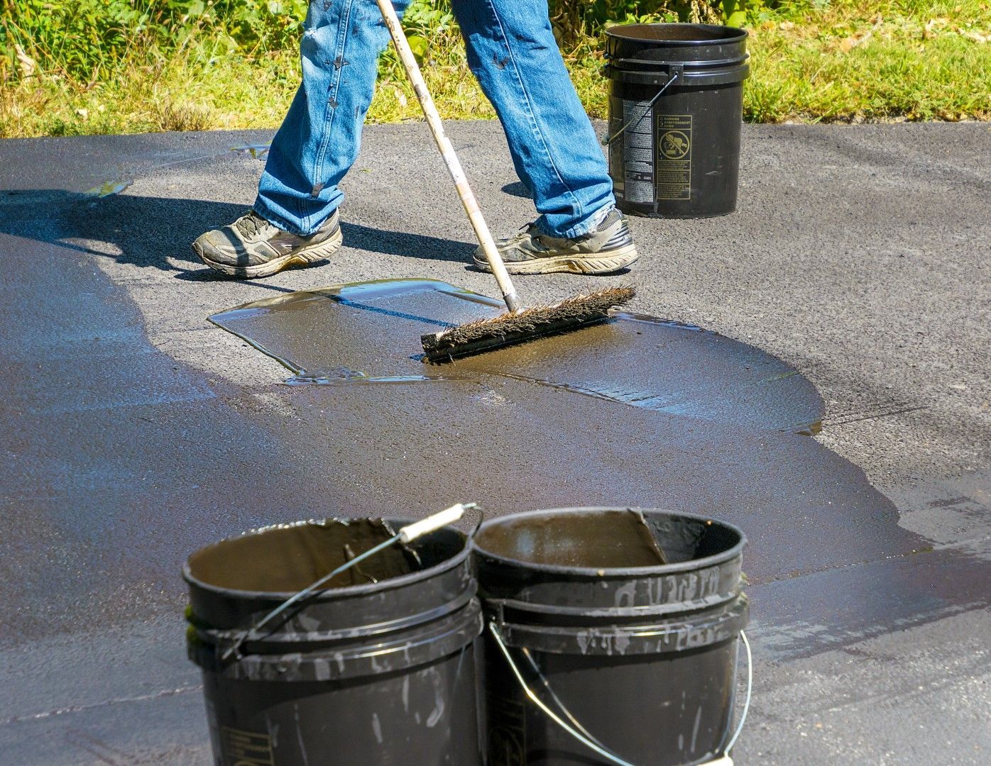 Person applying asphalt sealant to driveway with a brush, next to buckets of sealant.