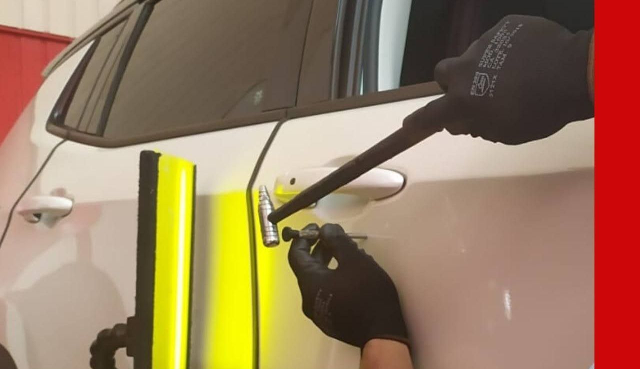 Pessoa usando uma ferramenta para trabalhar na porta branca de um carro perto de um painel refletor amarelo brilhante.