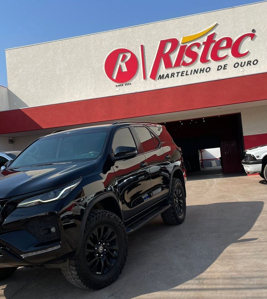 SUV preto estacionado em frente a uma concessionária Ristec com uma placa vermelha e branca acima da entrada.