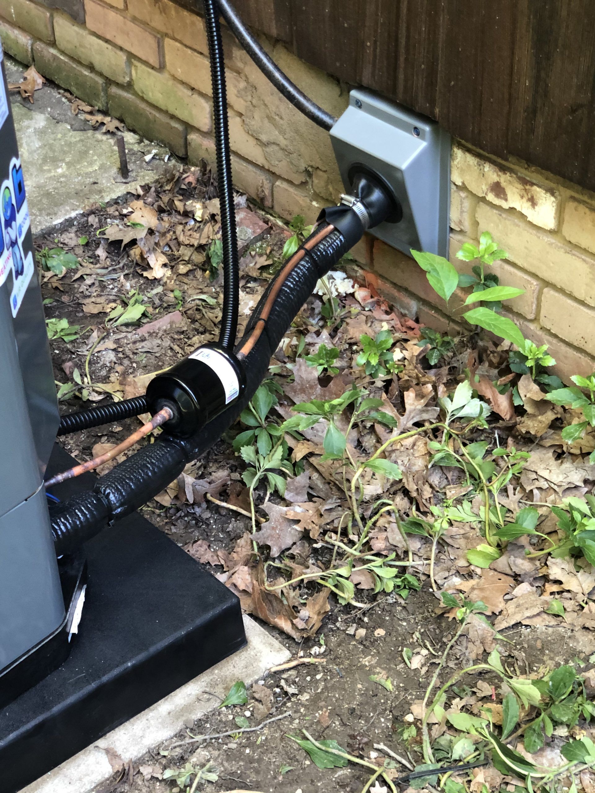 AC unit with black tubes and electrical connection next to brick wall and foliage.
