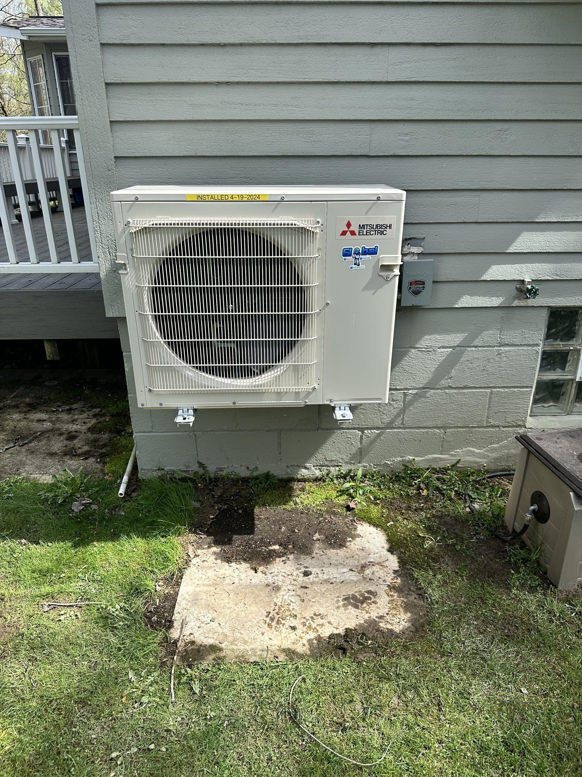 Mitsubishi air conditioning unit mounted on a gray house exterior, next to a deck and small concrete pad.
