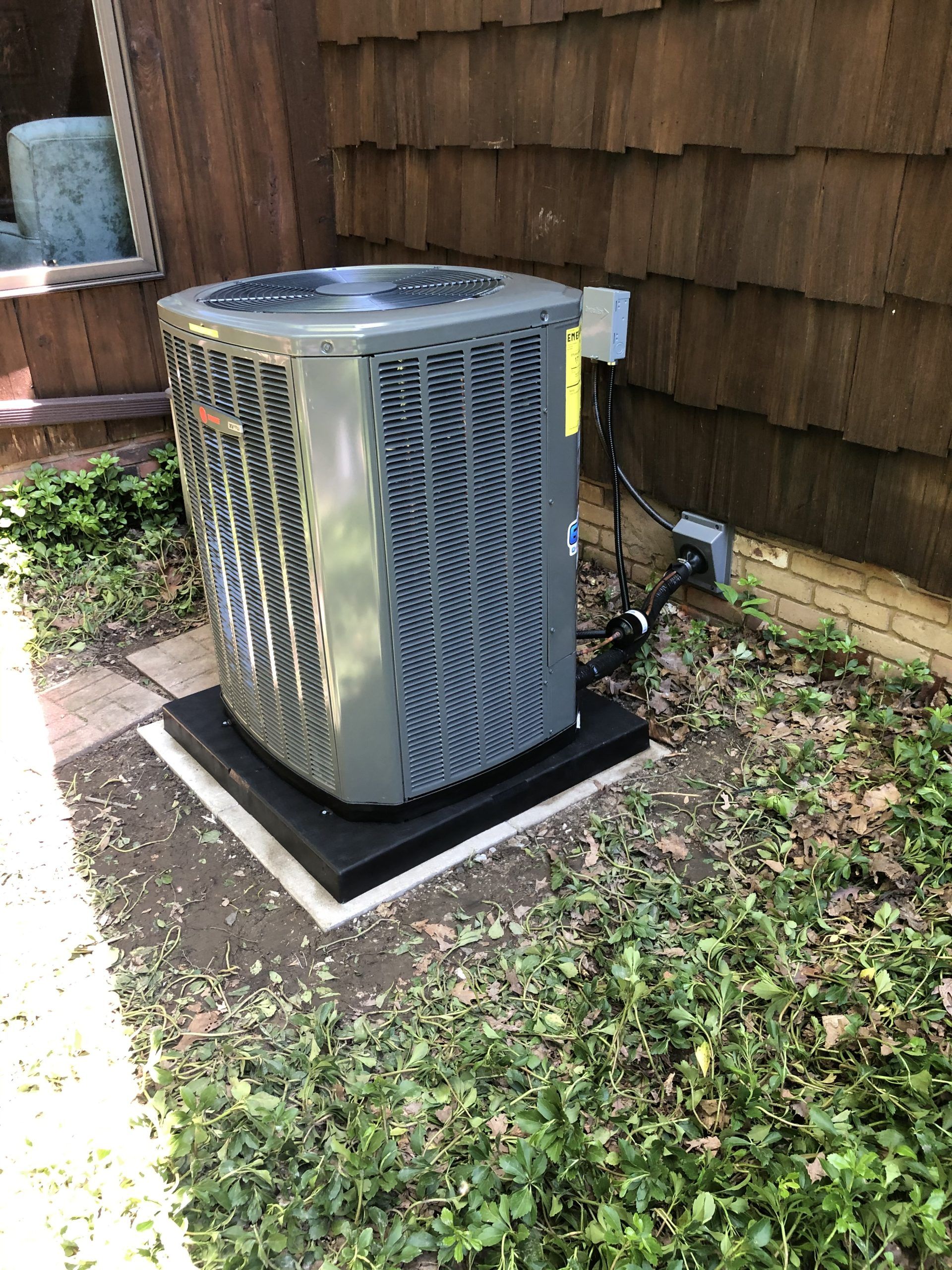 Air conditioning unit outside a brown-shingled building, sitting on a black base surrounded by shrubs and ground cover.