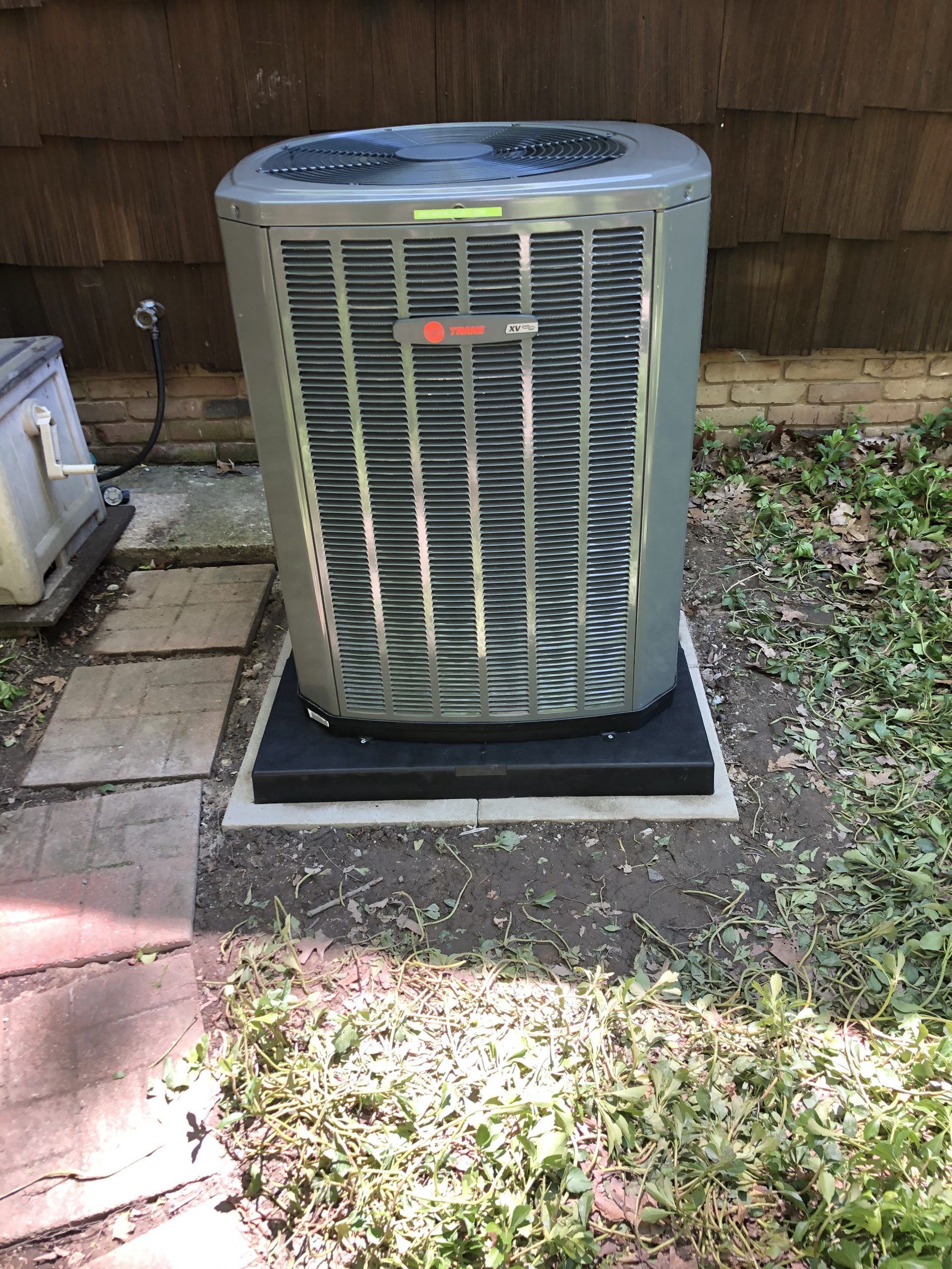 An air conditioning unit on a black platform, surrounded by grass and pavers. A wood-sided building is in the background.