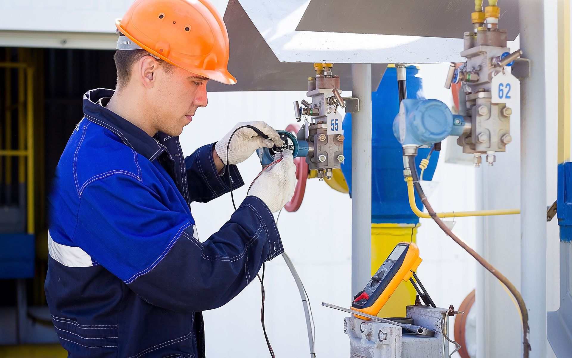 Man in work clothes and orange hard hat using a multimeter on industrial equipment outdoors.
