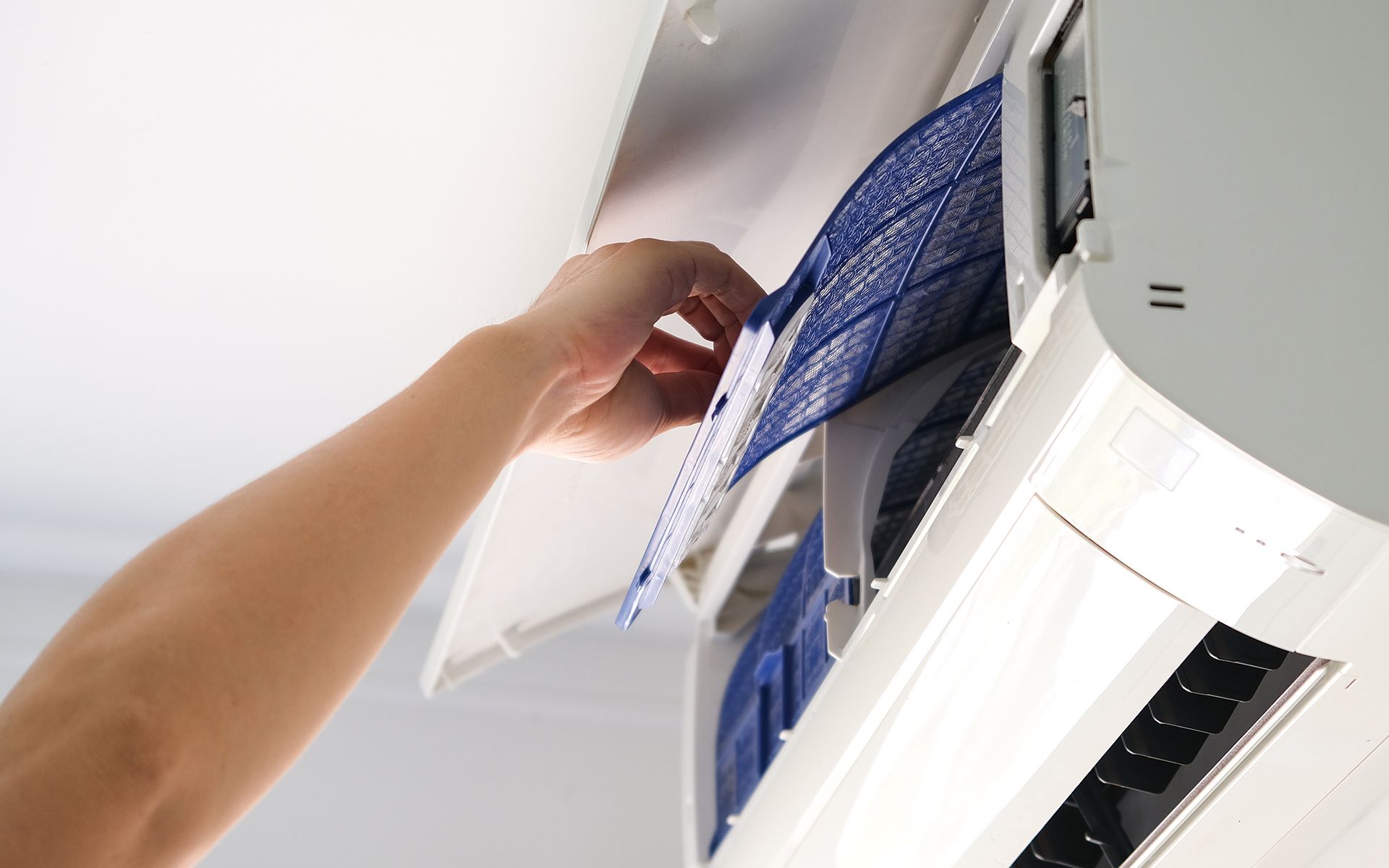 Hand removing an air filter from a white air conditioner on a white wall.