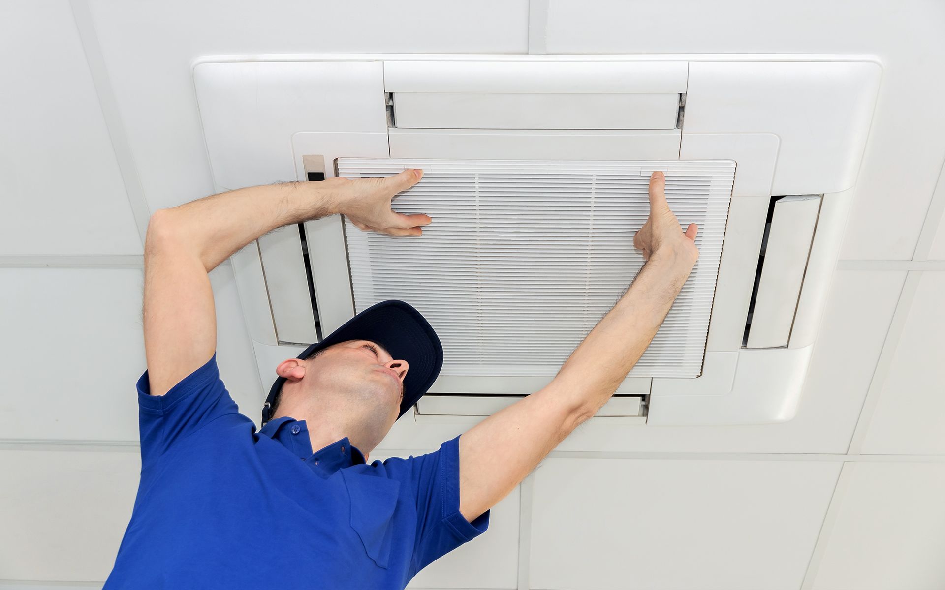 Man in blue shirt and cap inspecting a ceiling-mounted air vent, pointing at it.