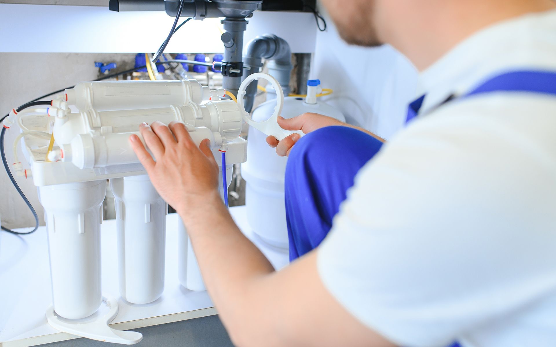 Plumber in white shirt and blue overalls, installing a water filter system under a sink.