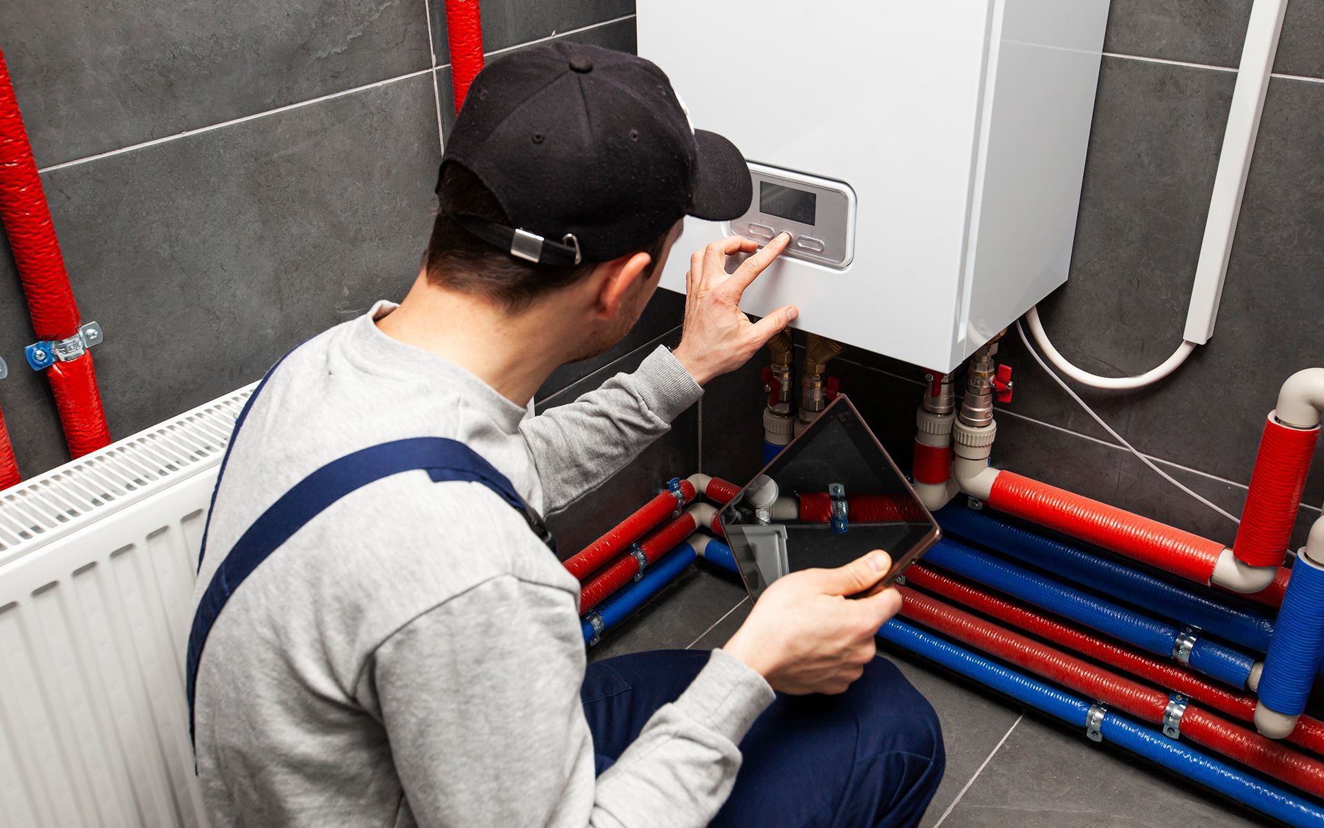 Plumber inspecting a boiler, holding a tablet. Red and blue pipes are visible against a gray wall.