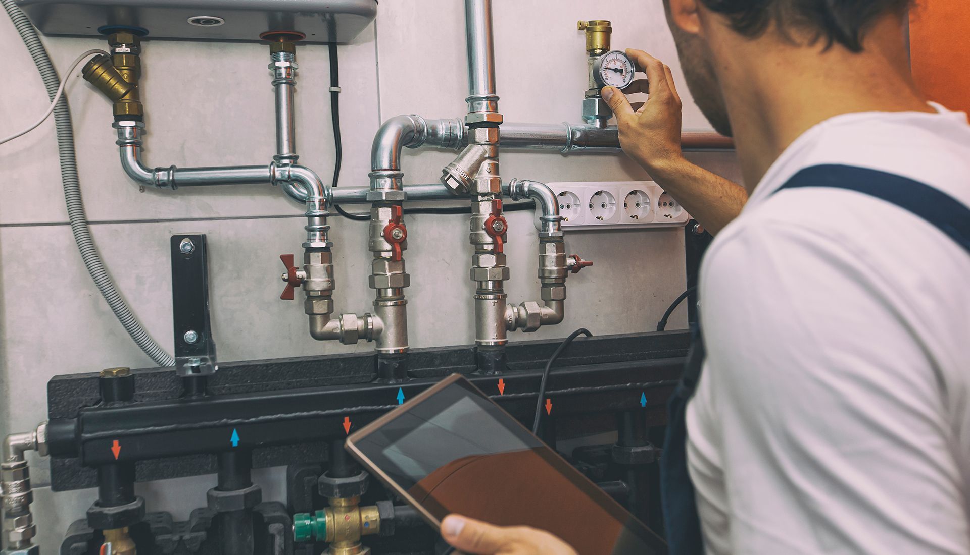 Plumber adjusting valves on pipes, using a tablet, in a utility room.