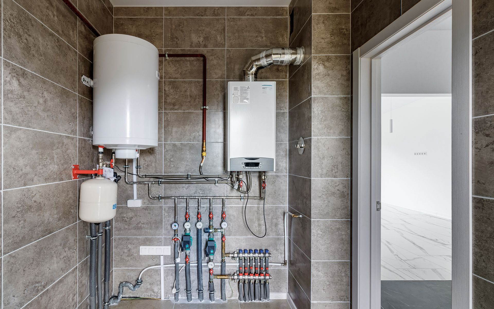 Water heater and boiler in a utility room with pipes and a door. Gray tiled wall.