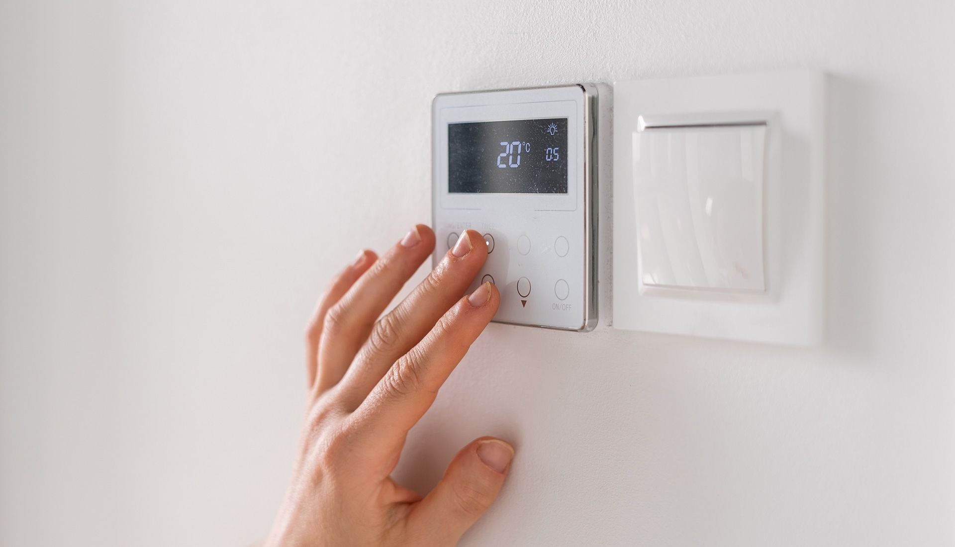Hand adjusting a digital thermostat on a white wall; the display shows 20 degrees Celsius.