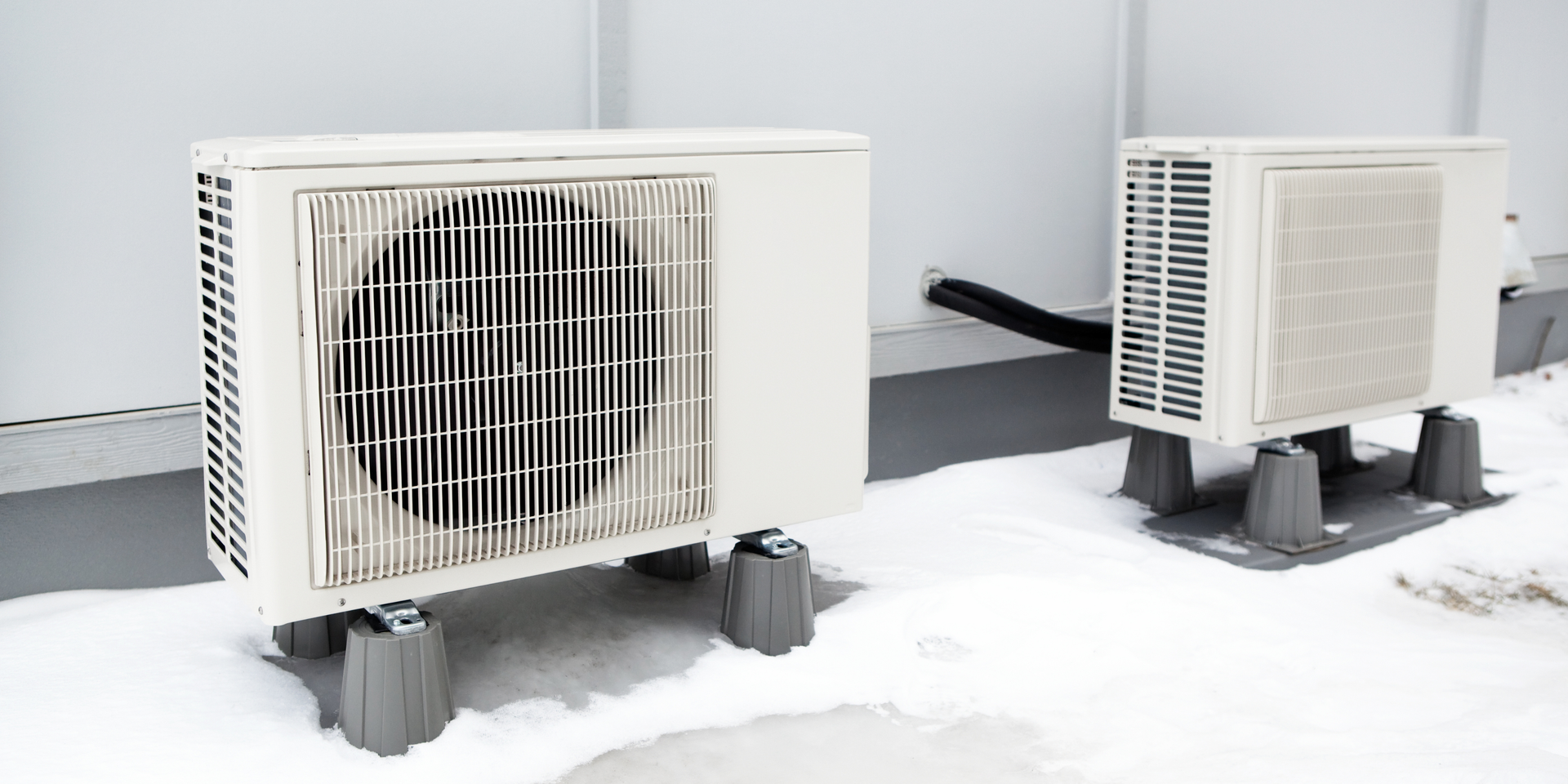 Two outdoor HVAC units elevated on gray blocks in snow against a white wall