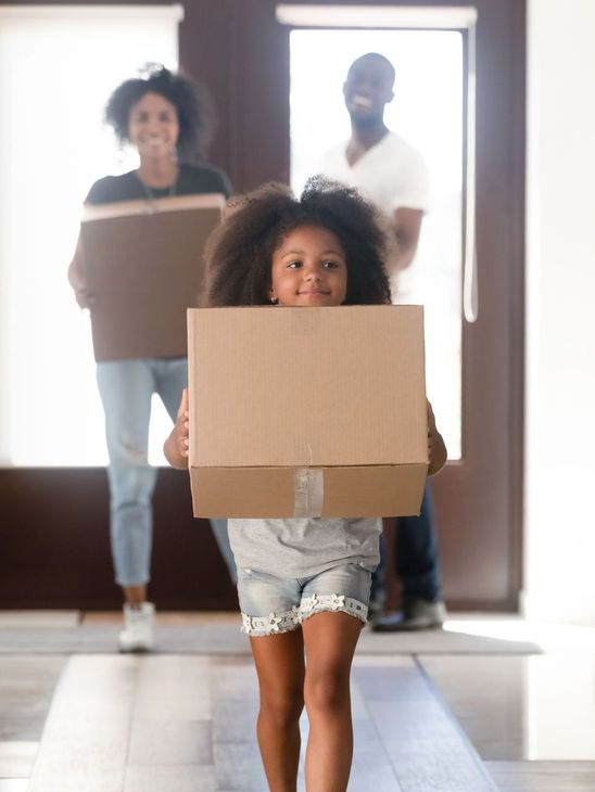 Family moving into a new home. Little girl carrying a box, parents behind her.